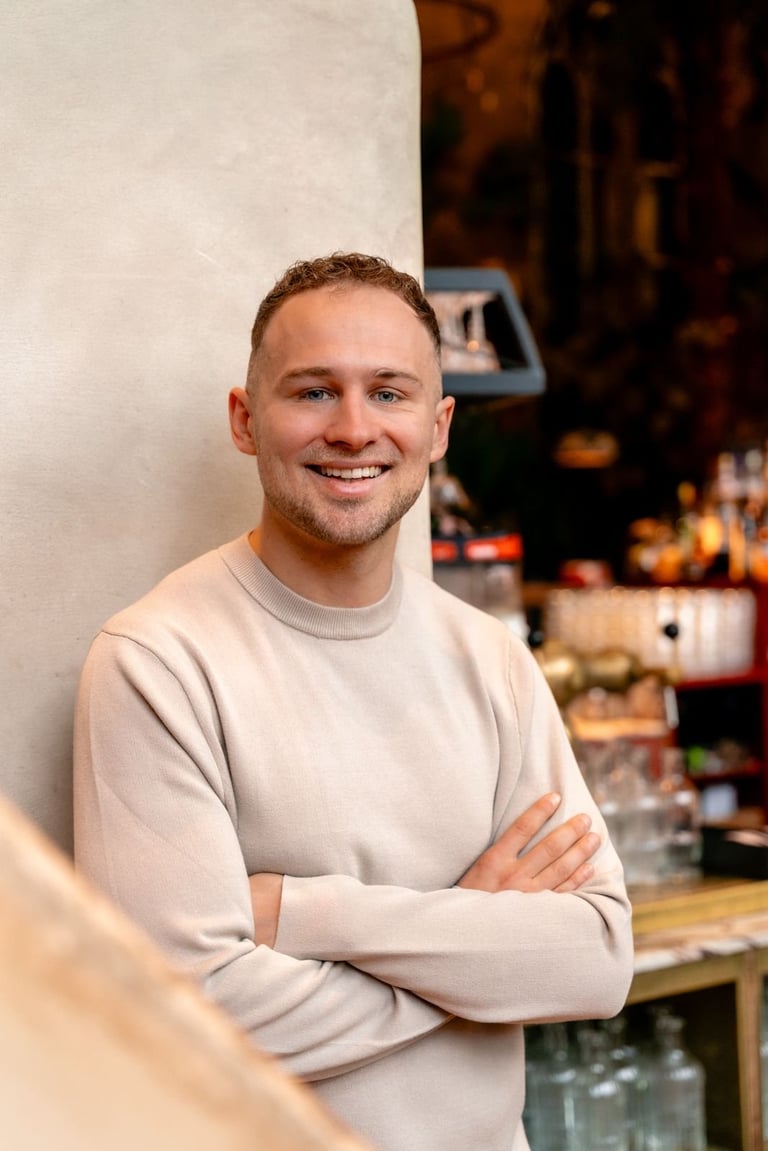 Smiling man with crossed arms wearing a beige sweatshirt, standing indoors against a white wall